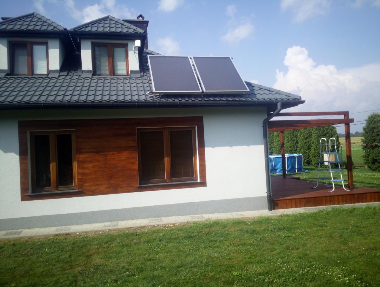 Nowoczesny dom jednorodzinny z panelami solarnymi na dachu i drewnianą elewacją, widoczny taras z basenem w tle, jasne niebo.