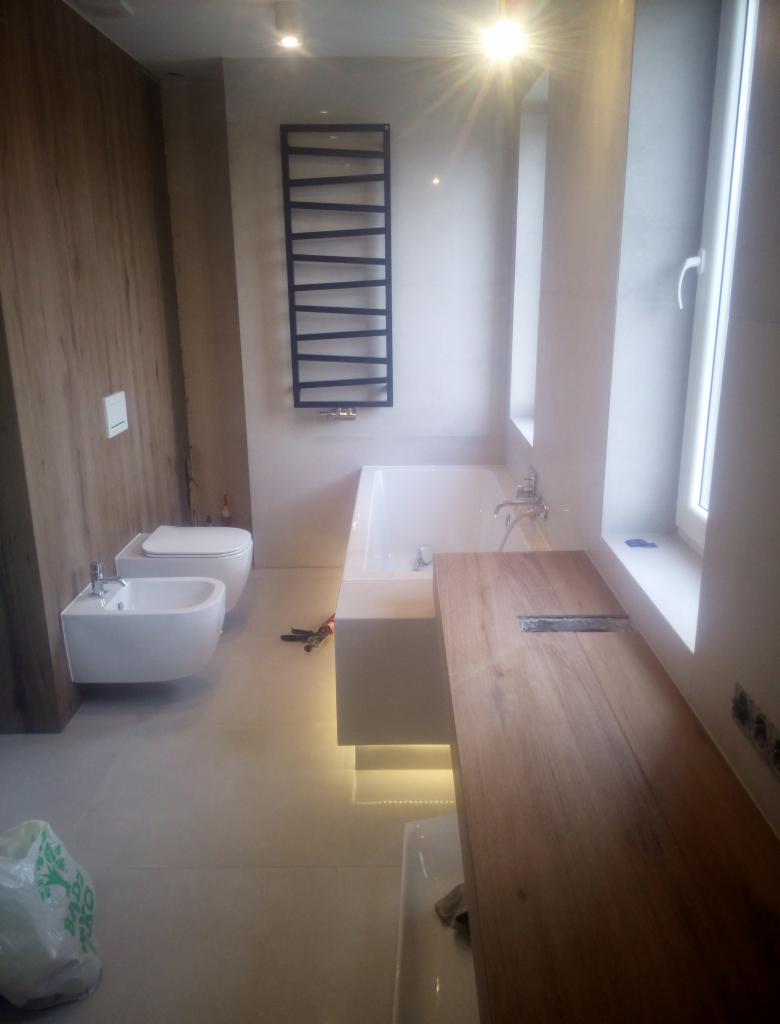 Nowoczesna łazienka z drewnianym blatem, białą armaturą i grzejnikiem drabinkowym, widoczne narzędzia na podłodze.