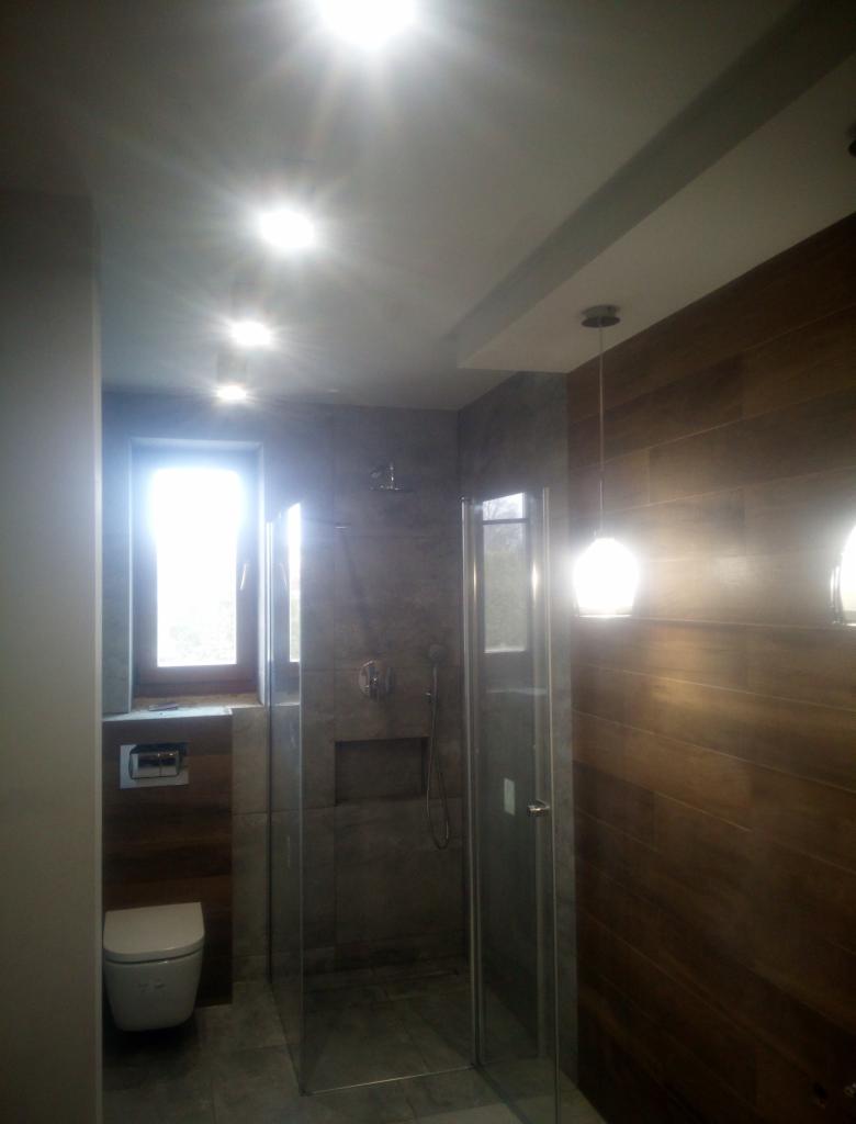 Nowoczesna łazienka z kabiną prysznicową typu walk-in, wykończona płytkami imitującymi drewno i szarym kamieniem, z wiszącą toaletą i minimalistycznym oświetleniem sufitowym.