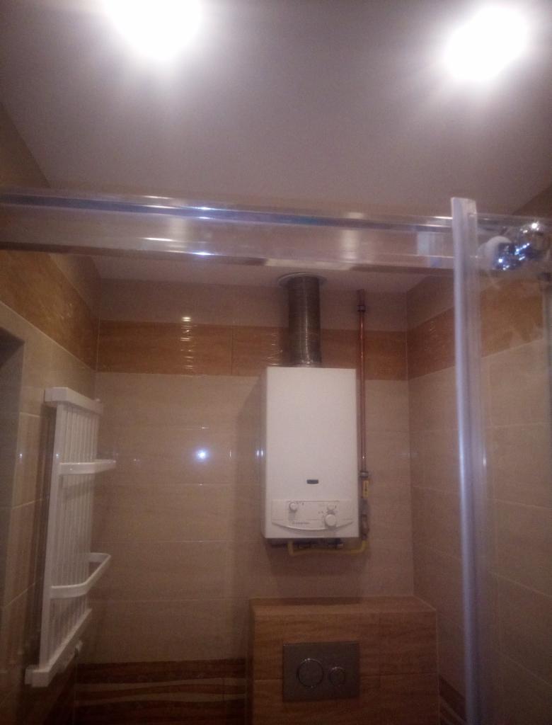 Biały gazowy podgrzewacz wody zamontowany na kafelkowanej ścianie w łazience, widoczne rury doprowadzające gaz i odprowadzające spaliny, obok grzejnik drabinkowy i szklana kabina prysznicowa.