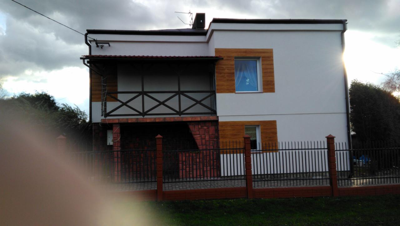 Dwupiętrowy dom z balkonem, wykończony białą elewacją i drewnopodobnymi panelami, z czarnym metalowym ogrodzeniem na ceglanej podmurówce, uchwycony w pochmurny dzień.