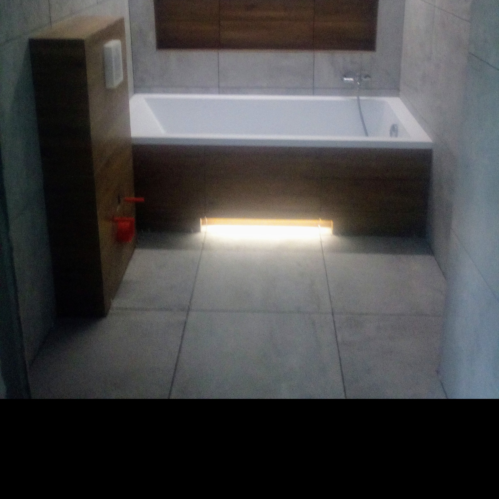 Nowoczesna łazienka z podświetlaną wanną, drewnianą zabudową i szarą, kwadratową płytką podłogową oraz zabudową geberit z czerwonymi elementami.