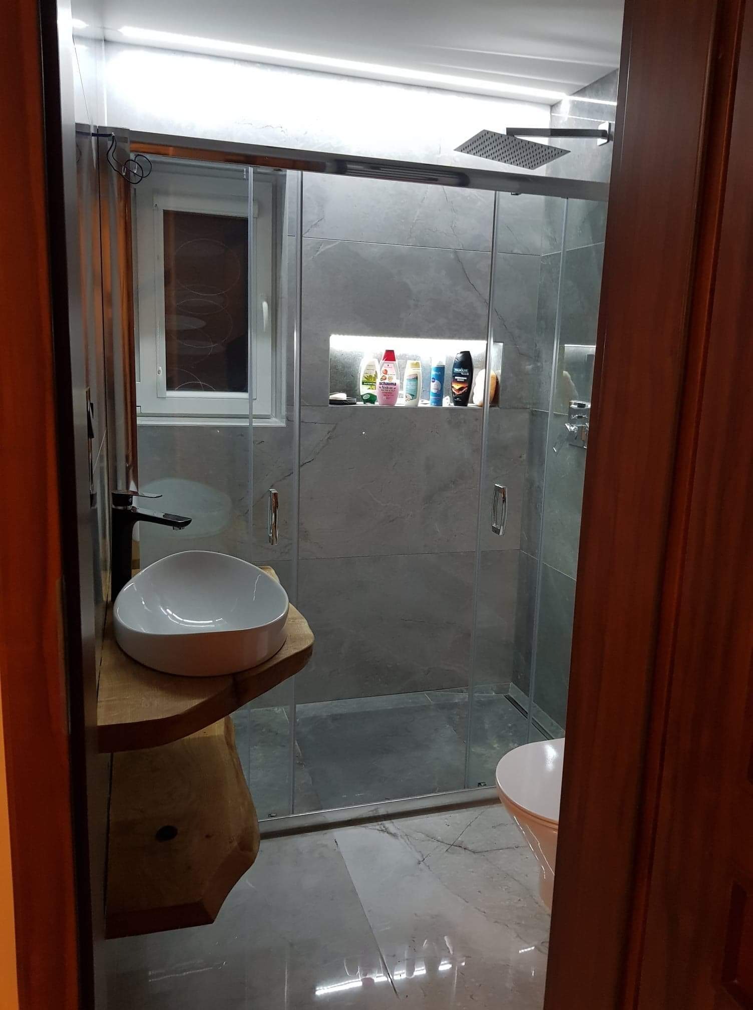 Nowoczesna łazienka z kabiną prysznicową typu walk-in, umywalką nablatową na drewnianym blacie i szarą glazurą imitującą marmur, oświetlona punktowo.