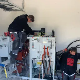 Dwóch elektryków pracuje przy rozdzielnicy elektrycznej ABB w jasnym pomieszczeniu. Jeden stoi na drabinie, drugi układa grube kable. Widoczne narzędzia i elementy instalacji.