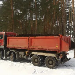 Czerwona ciężarówka MAN z otwartą skrzynią ładunkową wypełnioną ziemią, stoi na śniegu w otoczeniu wysokich drzew. Widoczny las w tle.