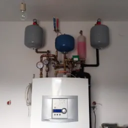 Biały piec grzewczy Junkers z podłączonymi zbiornikami wyrównawczymi i rurami miedzianymi na tle białej ściany, widoczna żarówka na suficie.