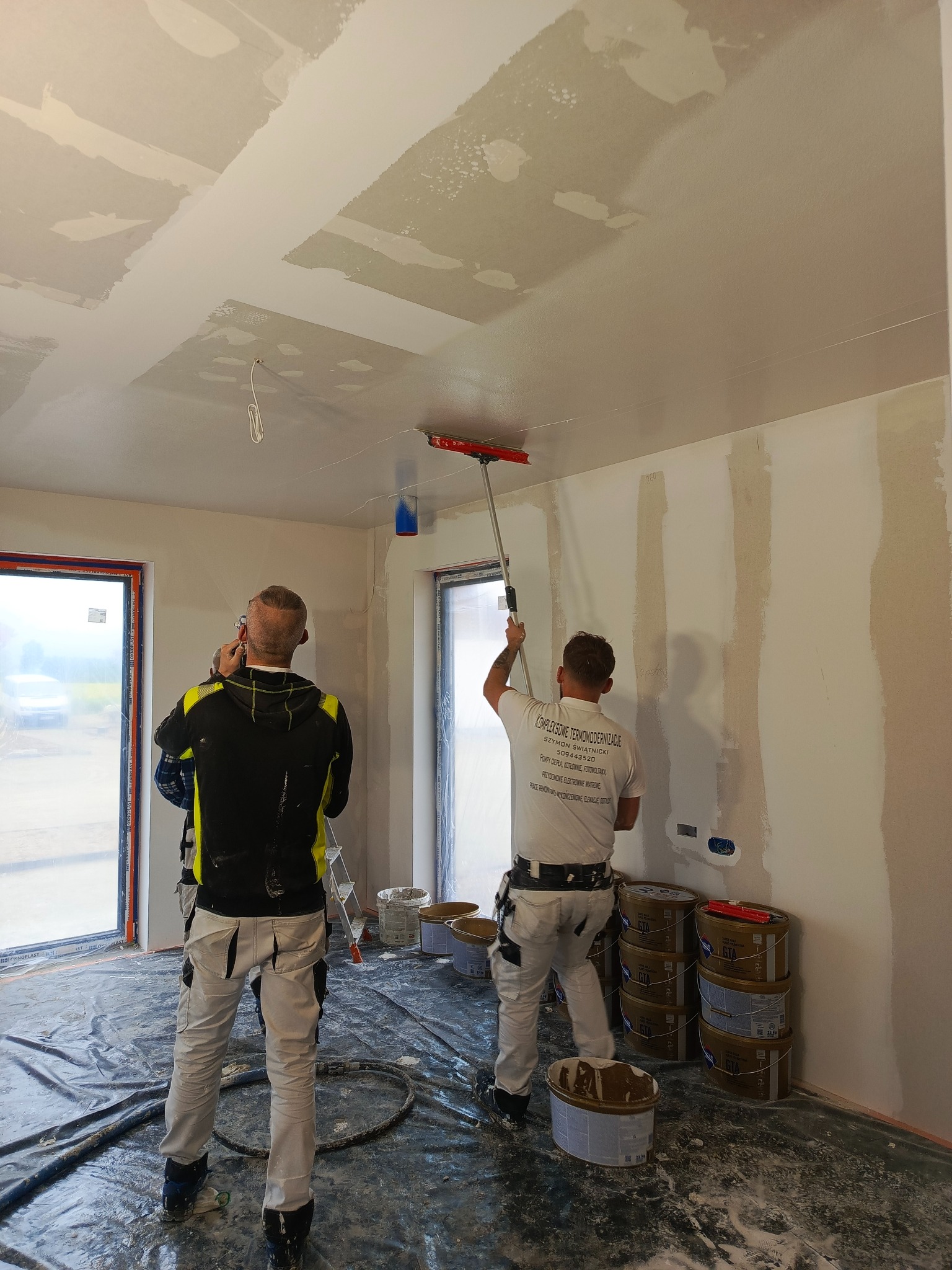Dwóch mężczyzn w trakcie szpachlowania ścian i sufitu w nowym wnętrzu. Jeden używa wałka na kiju, drugi robi zdjęcie. Folia ochronna na podłodze, widoczne puszki z gładzią.