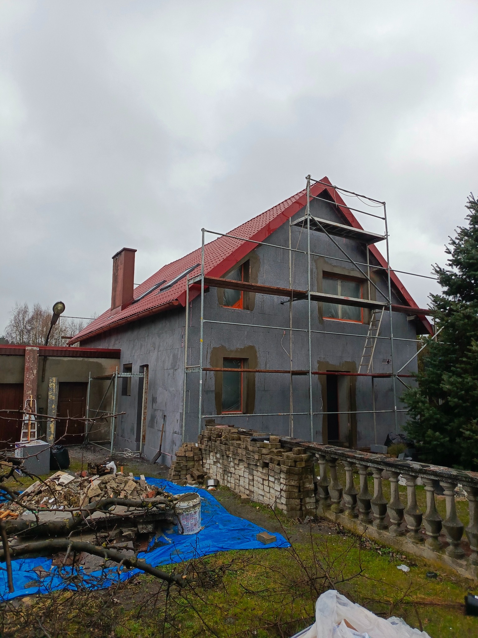 Dom z czerwonym dachem w trakcie ocieplania, otoczony rusztowaniem. Widoczny gruz i bałagan na posesji. Szara elewacja z widocznymi otworami okiennymi.