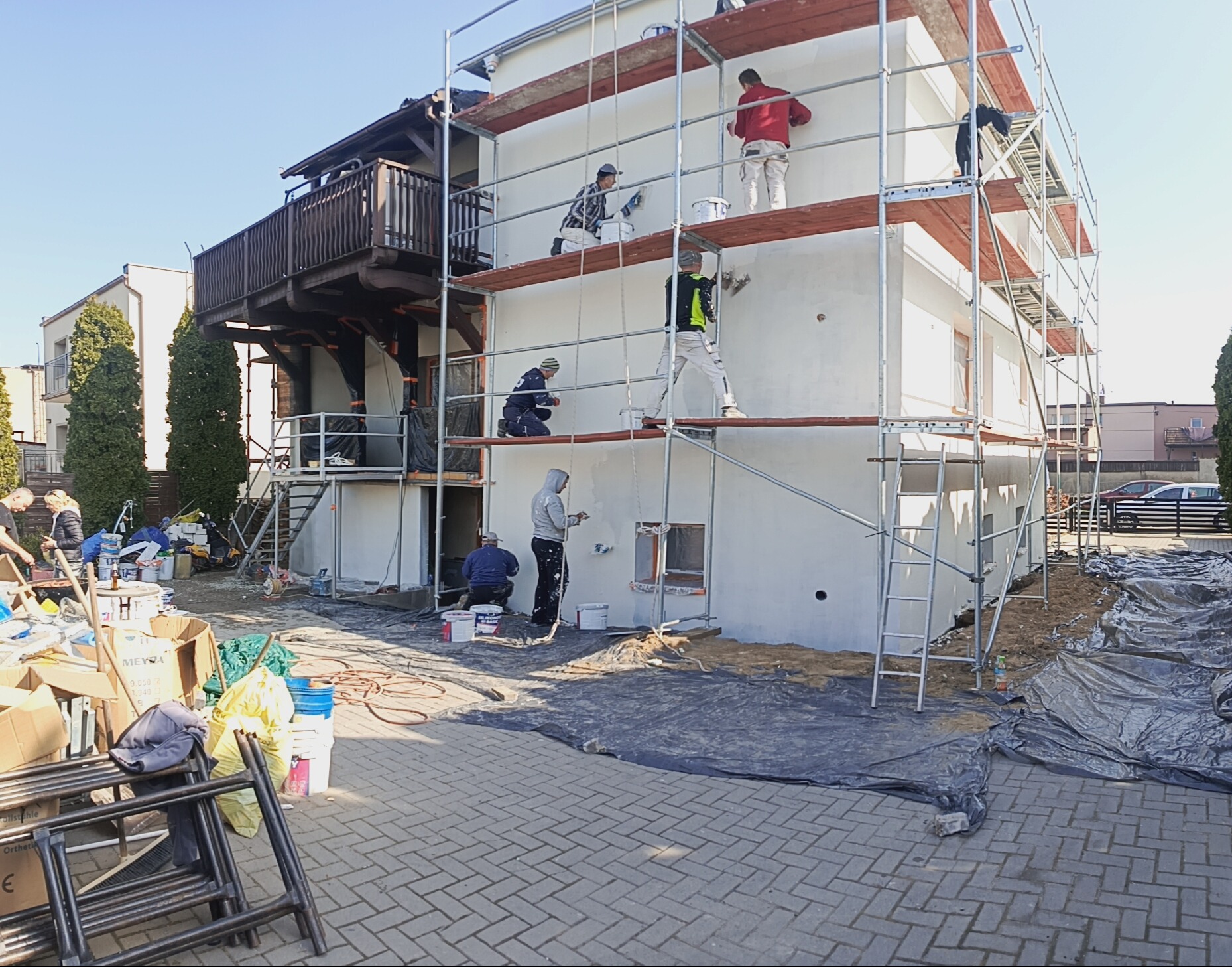 Ekipa malarzy na rusztowaniu odświeża elewację dwupiętrowego domu z drewnianym balkonem. Widoczne farby, narzędzia i zabezpieczenia wokół budynku.