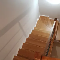 Wysoki kąt widzenia na drewniane schody z poręczą, widoczne słoje drewna i biała ściana w tle.