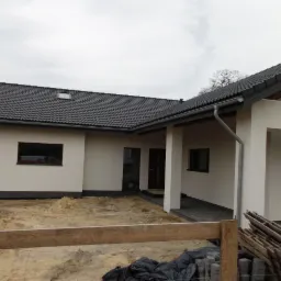 Nowoczesny dom jednorodzinny z garażem w trakcie wykańczania elewacji, widoczny fragment terenu przed budynkiem z piaskiem i materiałami budowlanymi.