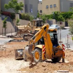 Żółty koparko-ładowarka i spychacz na placu budowy, wykop pod instalacje w miejskiej scenerii. Prace ziemne w trakcie realizacji, widoczne budynki w tle.