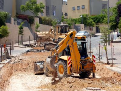 Żółty koparko-ładowarka i spychacz na placu budowy, wykop pod instalacje w miejskiej scenerii. Prace ziemne w trakcie realizacji, widoczne budynki w tle.