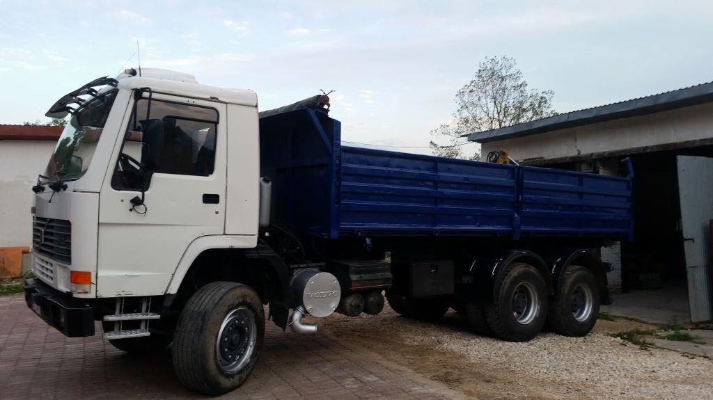 Biała ciężarówka z niebieską skrzynią ładunkową stoi na brukowanej powierzchni, obok budynku z otwartymi wrotami garażowymi, w tle drzewo i błękitne niebo.