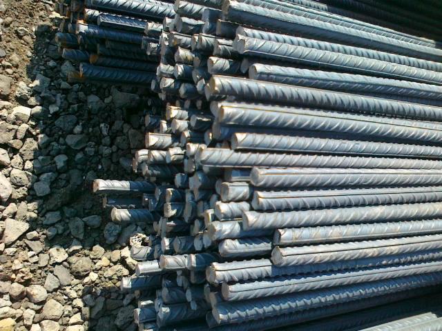 Stos żebrowanych prętów na tle ziemi i kamieni. Pręty ułożone równolegle, tworząc uporządkowany stos, widoczne żebrowania zwiększające przyczepność.