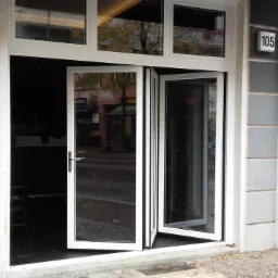 Białe składane okno aluminiowe z widocznym budynkiem po drugiej stronie ulicy, numer budynku 105 na ścianie obok.