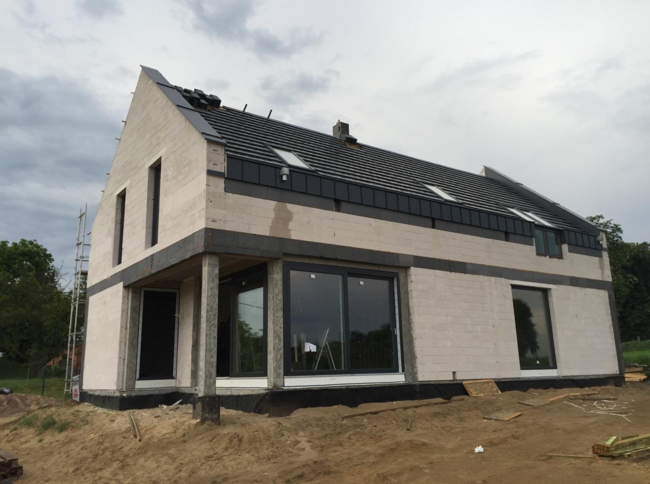 Dom jednorodzinny w trakcie budowy, widoczne duże przesuwne okna w czarnych ramach oraz fragment dachu z dachówką i oknami połaciowymi. Surowy stan elewacji z betonu komórkowego.