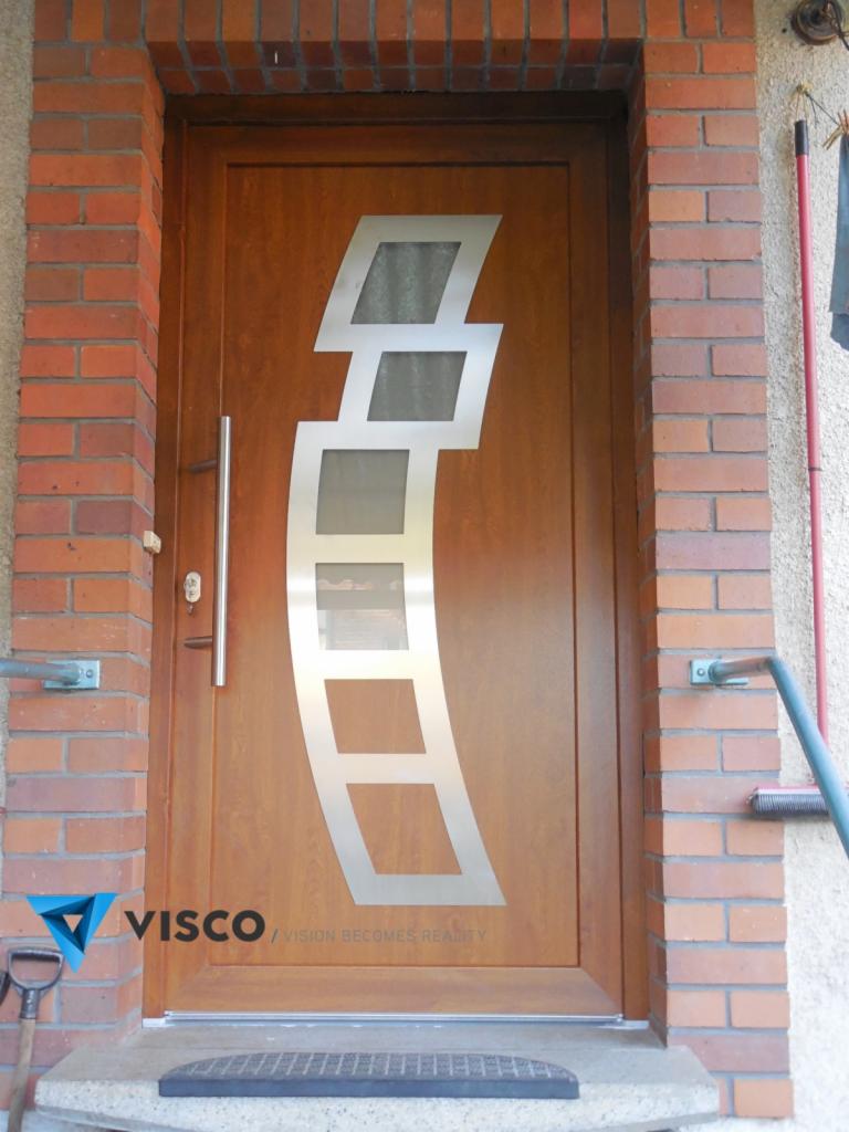 Brązowe drzwi wejściowe z nowoczesnym, geometrycznym zdobieniem ze stali nierdzewnej i przeszkleniami, umieszczone w murze z czerwonej cegły, z widocznym logo firmy w lewym dolnym rogu.