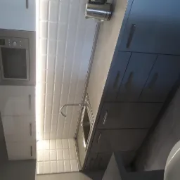 Nowoczesna kuchnia z szarymi szafkami, białymi płytkami ściennymi z delikatną fakturą i stalowym zlewem z baterią. Na blacie czajnik elektryczny. Ujęcie z dołu.