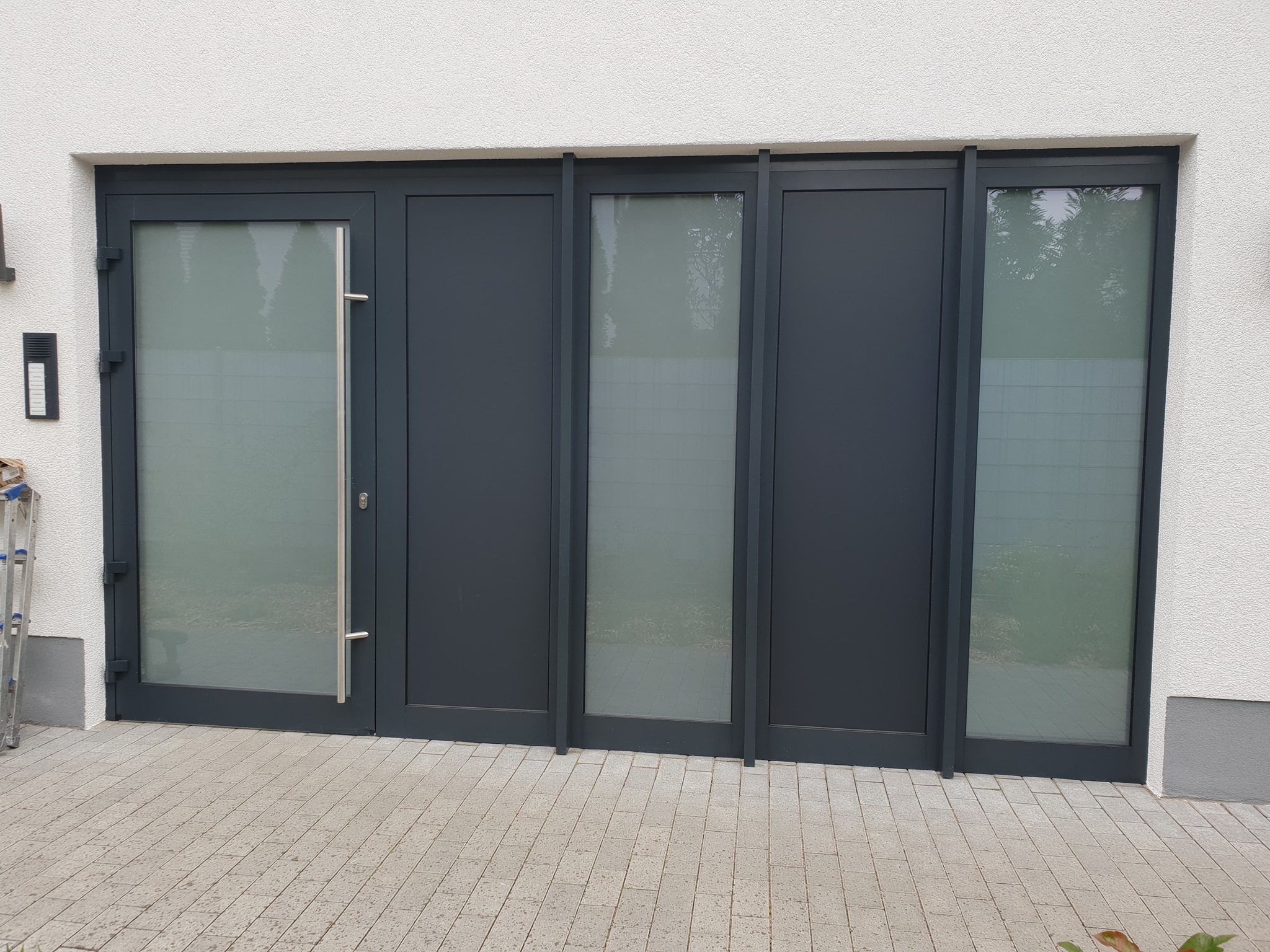 Nowoczesne, antracytowe drzwi przesuwne z matowego szkła, widoczne z zewnątrz, z drabiną opartą o ścianę po lewej stronie.