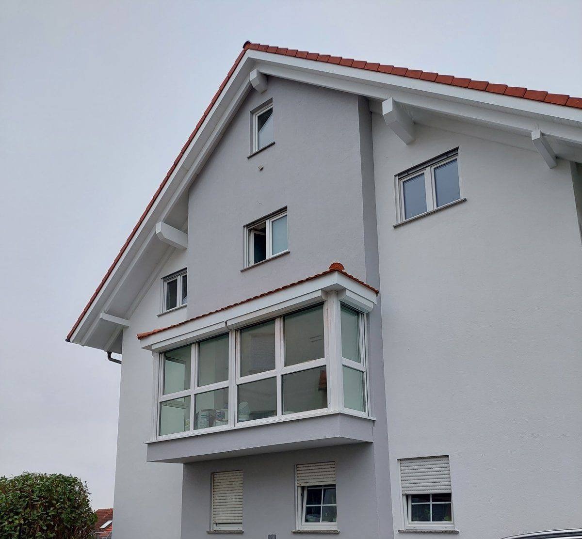 Szary budynek mieszkalny z białymi oknami PCV i roletami zewnętrznymi, widoczne z zewnątrz, z przeszklonym balkonem i czerwonym dachem.