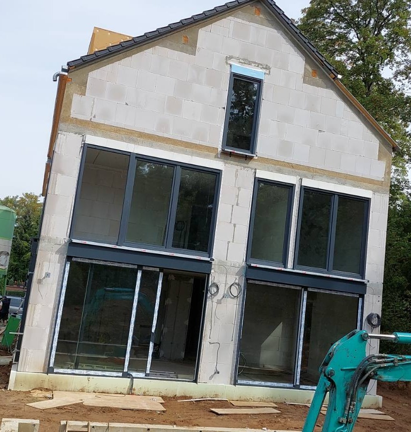 Budowa domu jednorodzinnego z zamontowanymi oknami i drzwiami przesuwnymi, widoczny stan surowy otwarty, z elementami wykończeniowymi i sprzętem budowlanym na pierwszym planie.