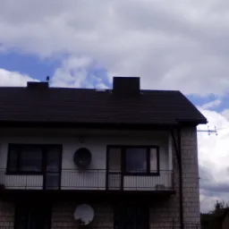 Dwukondygnacyjny dom z ciemnym, spadzistym dachem i balkonem, na tle błękitnego nieba z białymi obłokami. Widoczne anteny satelitarne na elewacji.