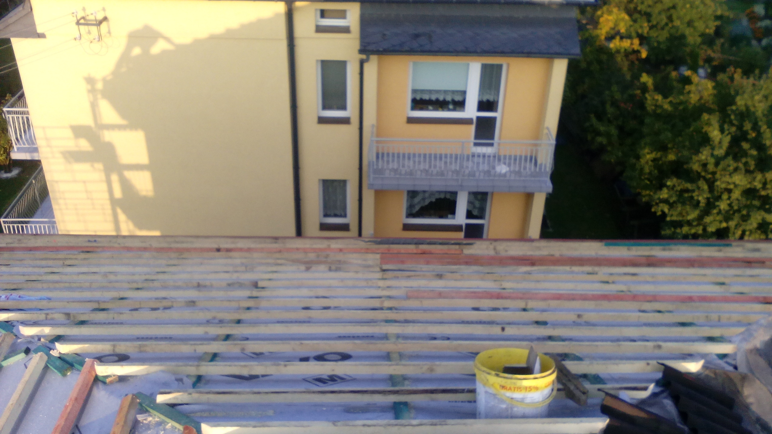 Wysoki kąt: konstrukcja dachu z łatami na folii, z żółtym wiadrem na pierwszym planie, budynek w tle. Widok na budynek z dachu w trakcie prac.