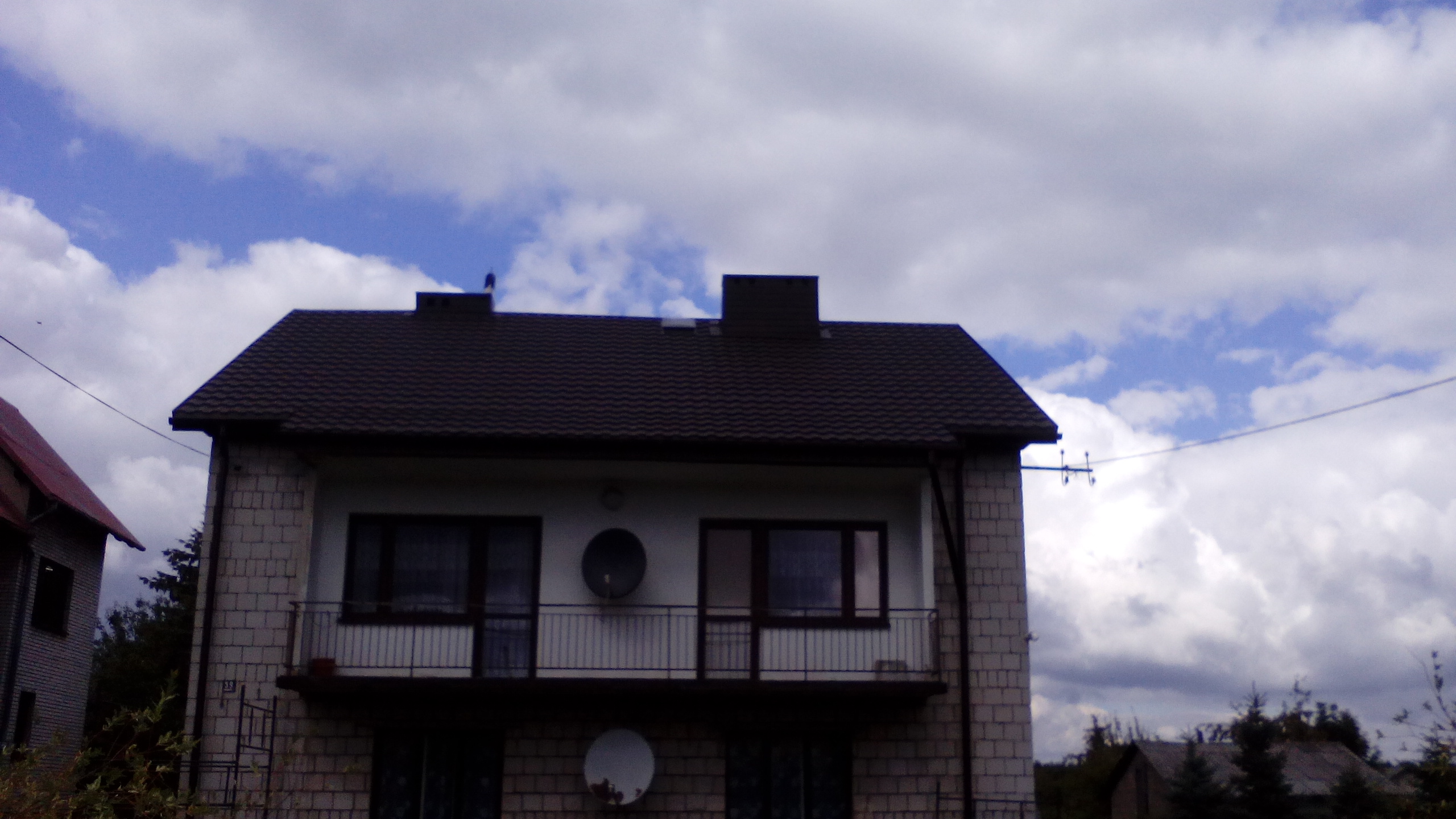Dwukondygnacyjny dom z ciemnym, spadzistym dachem i balkonem, na tle błękitnego nieba z białymi obłokami. Widoczne anteny satelitarne na elewacji.