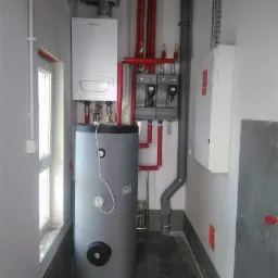 Kompaktowa kotłownia: biały piec gazowy kondensacyjny wiszący nad szarym zasobnikiem ciepłej wody, połączone czerwoną instalacją hydrauliczną, widoczne zabezpieczenia elektryczne i piony wentylacyjne.