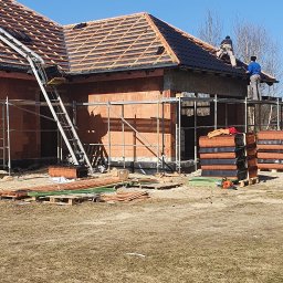 M&M BLACHARSTWO-DEKARSTWO MAREK MALISKA - Dwóch dekarzy na rusztowaniu układa dachówkę na nowym dachu domu jednorodzinnego, widoczny podnośnik do dachówki i palety z dachówką.