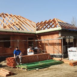 M&M BLACHARSTWO-DEKARSTWO MAREK MALISKA - Budowa domu jednorodzinnego w trakcie wznoszenia więźby dachowej; pracownik w niebieskiej bluzie siedzi na stosie drewnianych belek na tle częściowo pokrytego dachu i rusztowań.