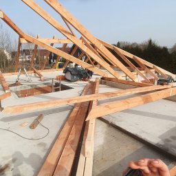 M&M BLACHARSTWO-DEKARSTWO MAREK MALISKA - Montaż drewnianej konstrukcji dachu w surowym stanie, z widocznymi elementami więźby, ceglanym kominem i ręką trzymającą telefon, fotografującą postęp prac.