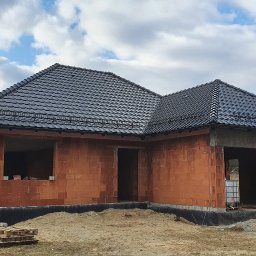 M&M BLACHARSTWO-DEKARSTWO MAREK MALISKA - Dom jednorodzinny w trakcie budowy z częściowo ukończonym, ciemnoszarym dachem z dachówki ceramicznej; widoczne ceglane ściany i przygotowany teren wokół.