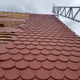 Układanie czerwonej dachówki karpiówki na drewnianej konstrukcji dachu, widoczna drabina dekarska i pochmurne niebo.