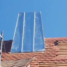 Montaż obróbki blacharskiej komina na dachu z czerwonej dachówki karpiówki, widoczne rusztowanie i błękitne niebo.