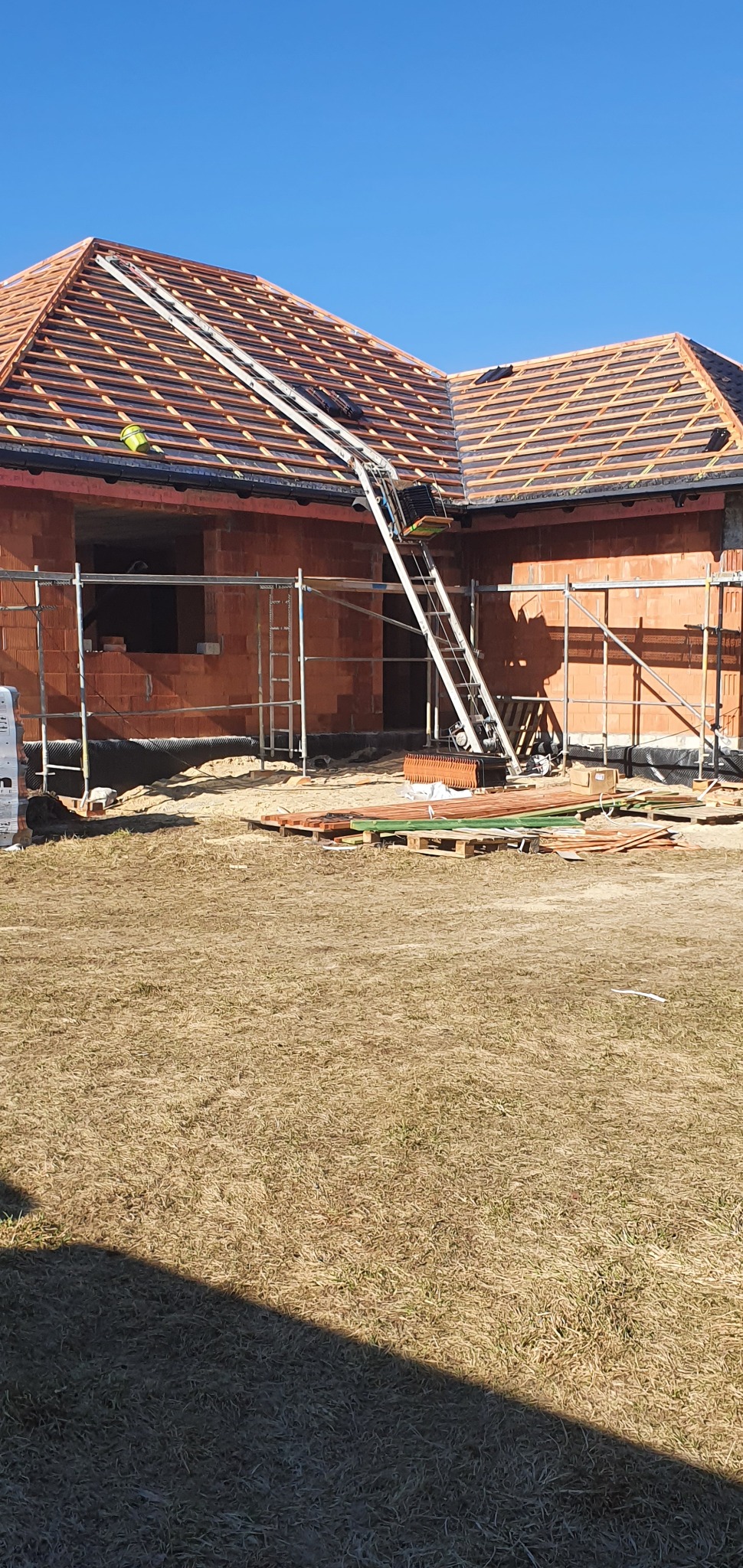 Budowa domu jednorodzinnego w trakcie krycia dachu dachówką ceramiczną, widoczny podnośnik do transportu materiałów budowlanych na dach.