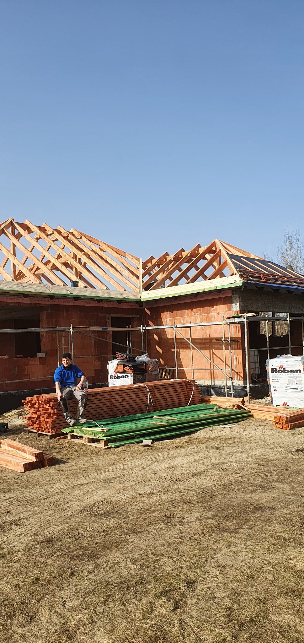 Budowa domu jednorodzinnego w trakcie wznoszenia więźby dachowej; pracownik w niebieskiej bluzie siedzi na stosie drewnianych belek na tle częściowo pokrytego dachu i rusztowań.