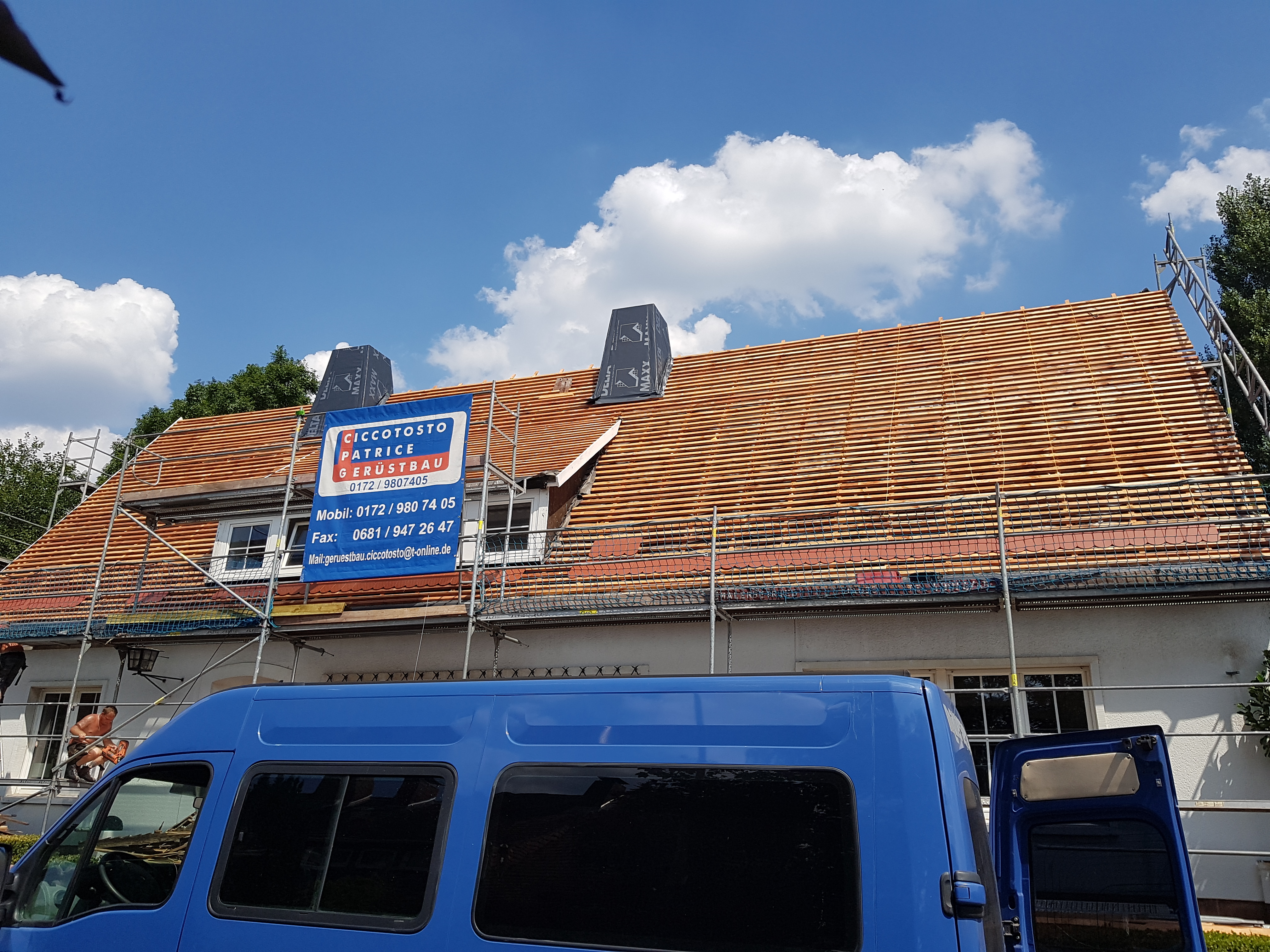 Dom jednorodzinny w trakcie układania drewnianej konstrukcji pod pokrycie dachowe, widoczne rusztowania i reklama firmy dekarskiej, na pierwszym planie niebieski samochód dostawczy.