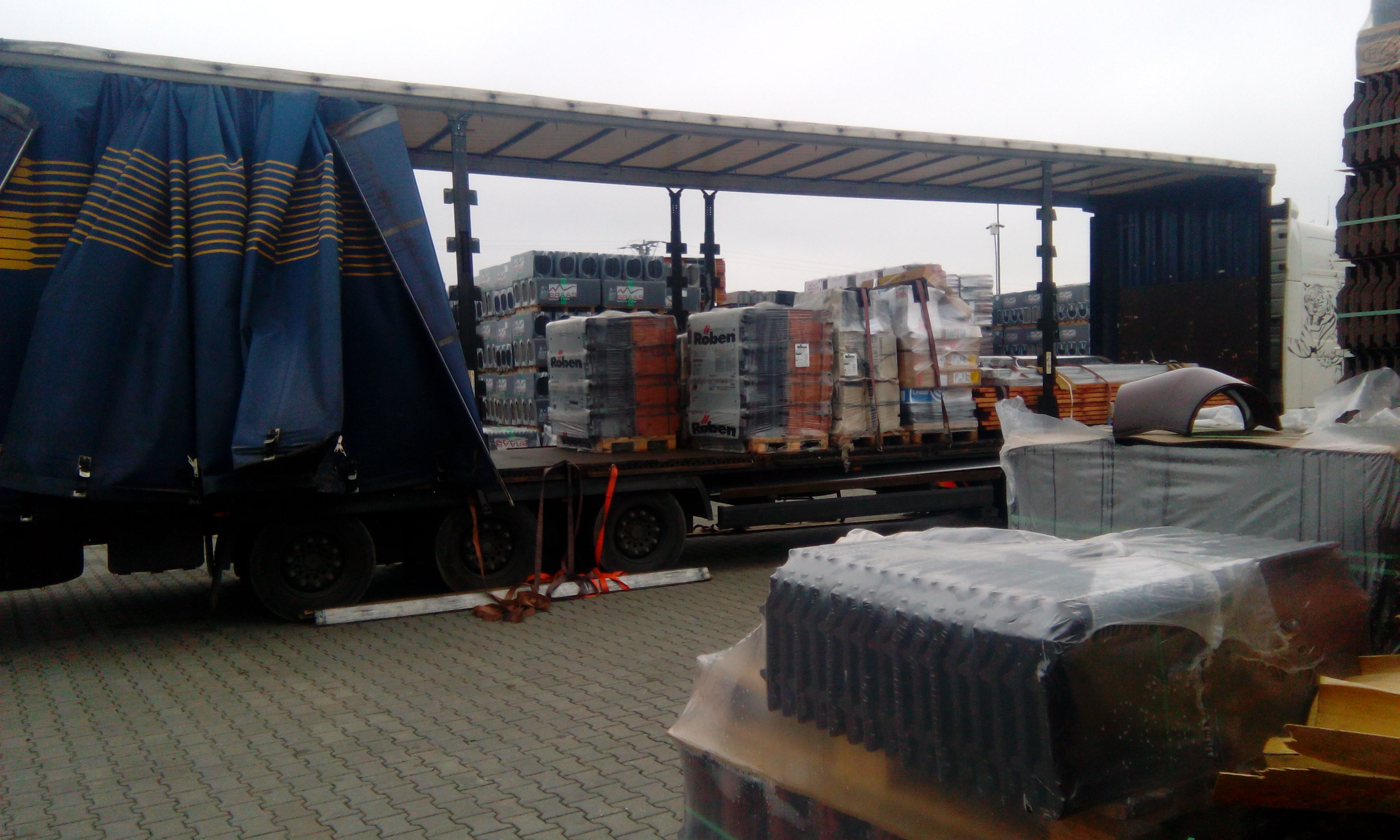 Ciężarówka z otwartą plandeką, załadowana paletami z dachówkami Roben, zaparkowana na brukowanym placu w pochmurny dzień.