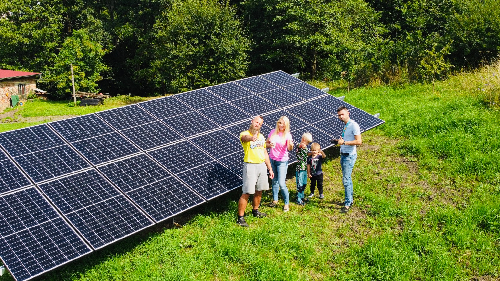 Rodzina pozuje przed nowo zainstalowaną instalacją fotowoltaiczną na trawiastym terenie z budynkiem gospodarczym w tle, wszyscy pokazują kciuki w górę.