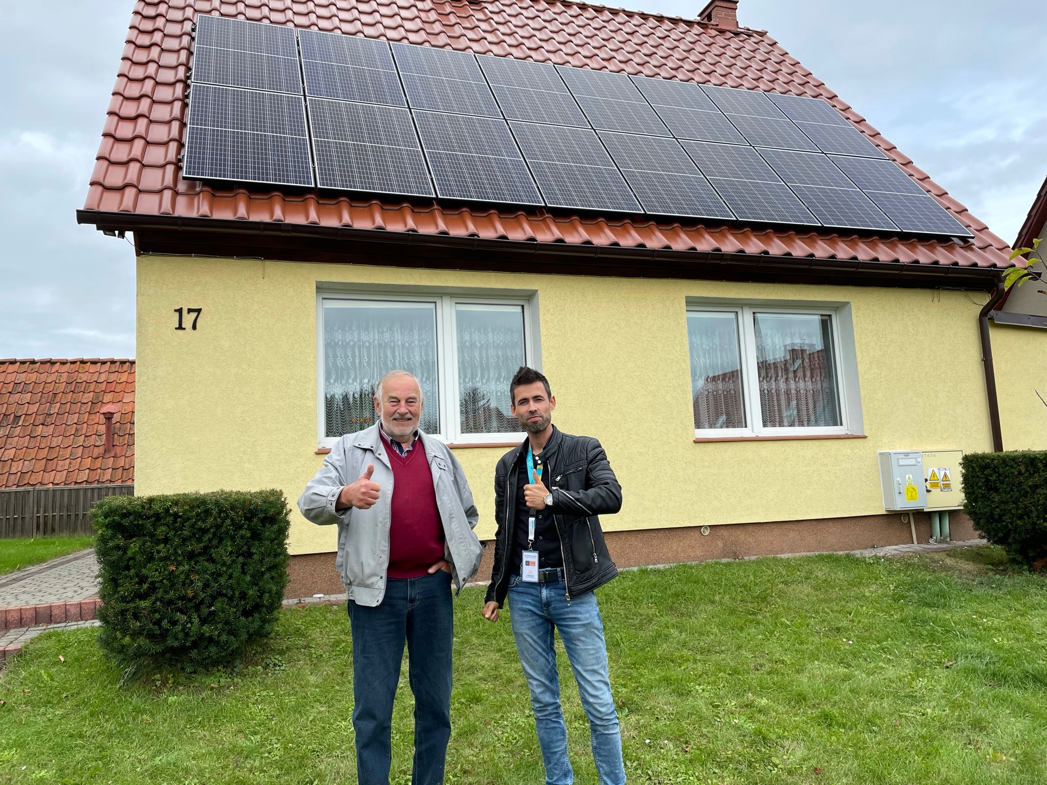 Dwaj mężczyźni pokazują kciuki w górę na tle domu z panelami słonecznymi na dachu z czerwonej dachówki, numer domu 17, trawnik i krzewy przed domem.