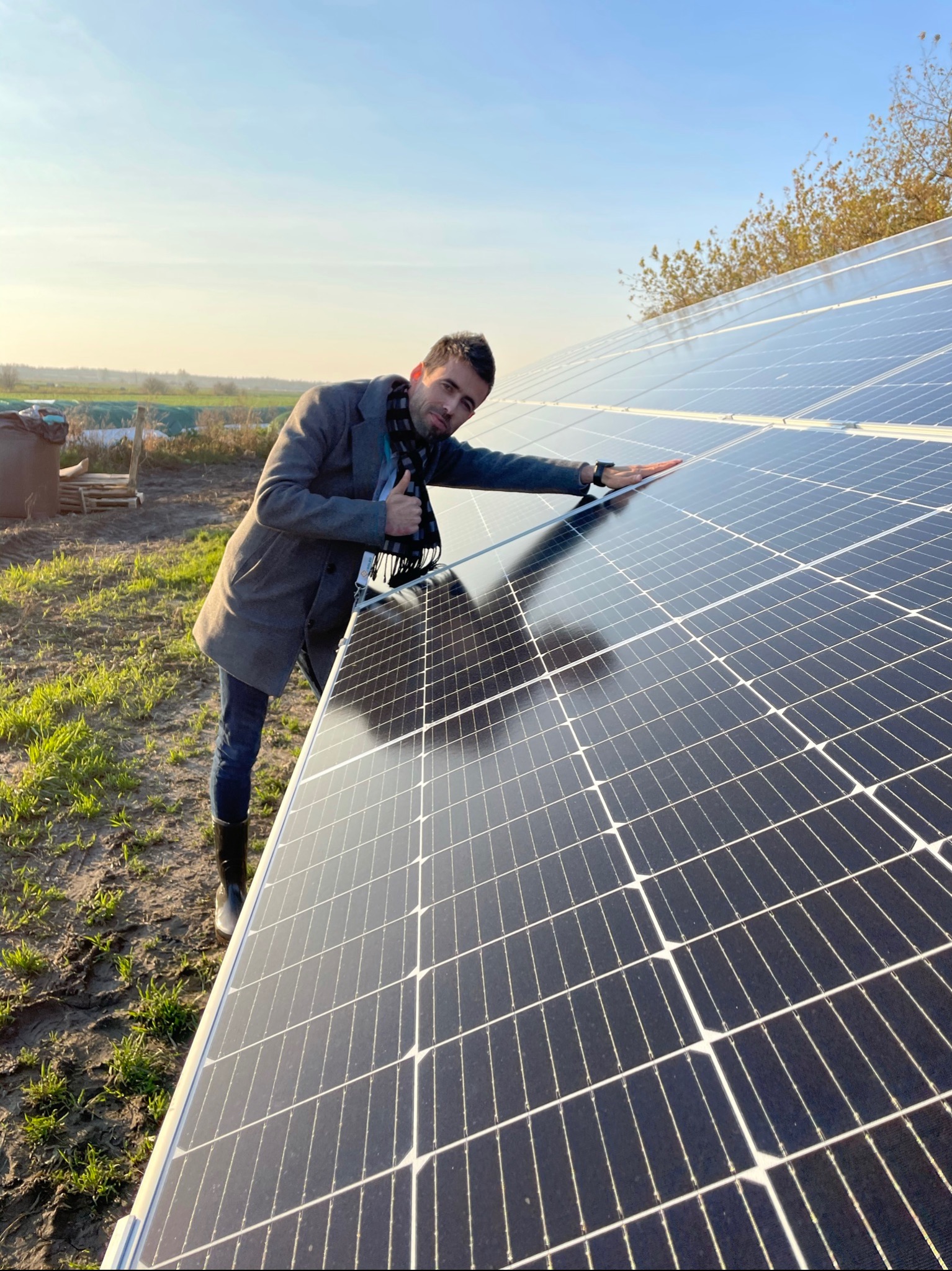 Mężczyzna w płaszczu dotyka paneli słonecznych, pokazując kciuk w górę na tle wiejskiego krajobrazu.