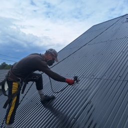 PHU Pro-Clean - Pracownik w uprzęży, z maską ochronną i pistoletem natryskowym, aplikuje powłokę na blaszany dach w pochmurny dzień, widoczny fragment budynku mieszkalnego w tle.
