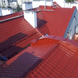 Pracownik w kasku, z narzędziami na dachu z czerwonej dachówki, widok z góry na budynki mieszkalne w tle.