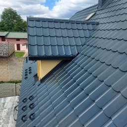 PHU Pro-Clean - Nowy, grafitowy dach z blachodachówki modułowej z kominem w kolorze dachu i oknem dachowym, widok z góry na tle błękitnego nieba z chmurami.