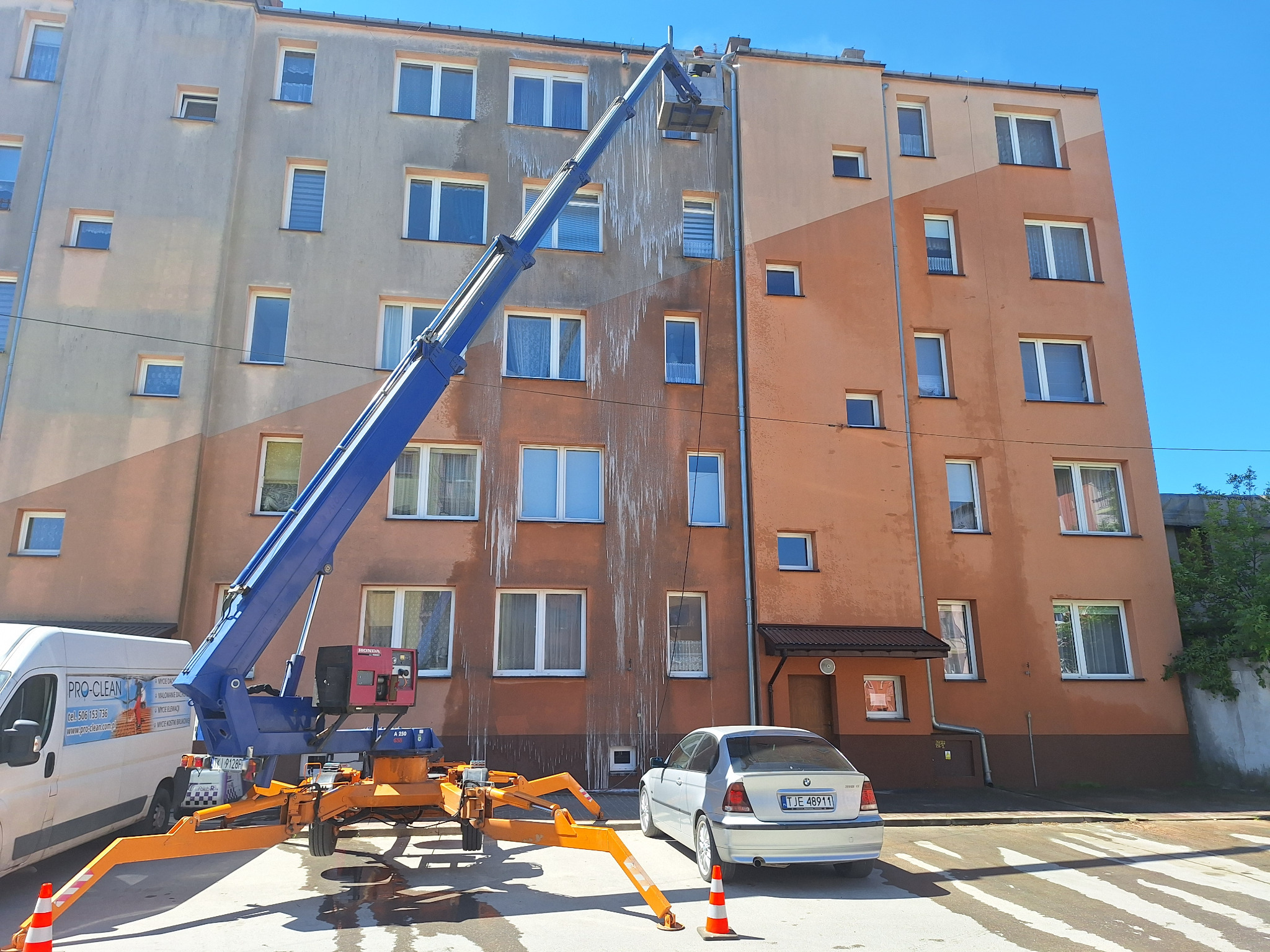 Pracownik na podnośniku koszowym czyści elewację wielopiętrowego budynku, widoczne zacieki po spływającej wodzie, na dole zaparkowany samochód i van firmy Pro-Clean z reklamą usług.