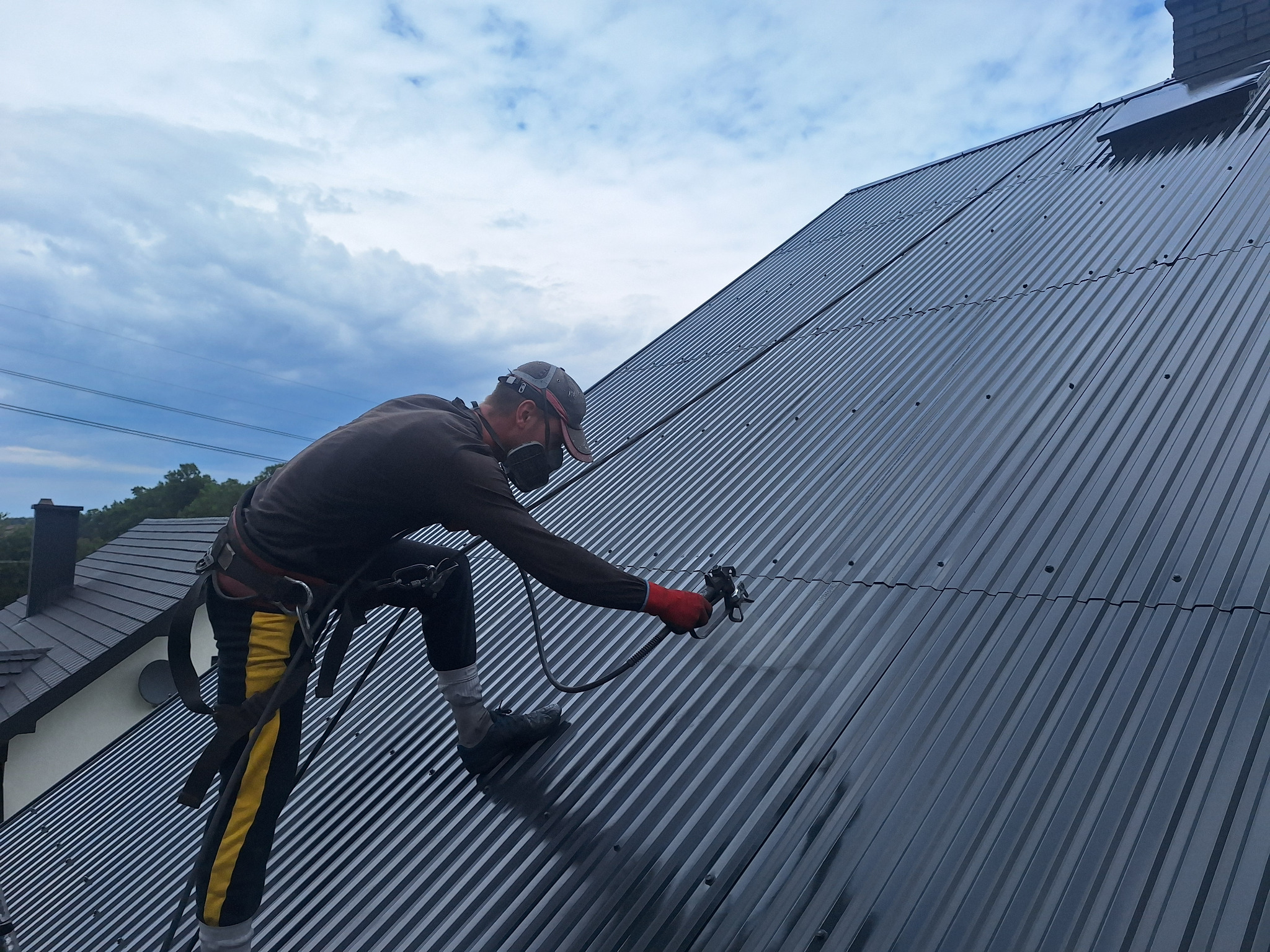 Pracownik w uprzęży, z maską ochronną i pistoletem natryskowym, aplikuje powłokę na blaszany dach w pochmurny dzień, widoczny fragment budynku mieszkalnego w tle.