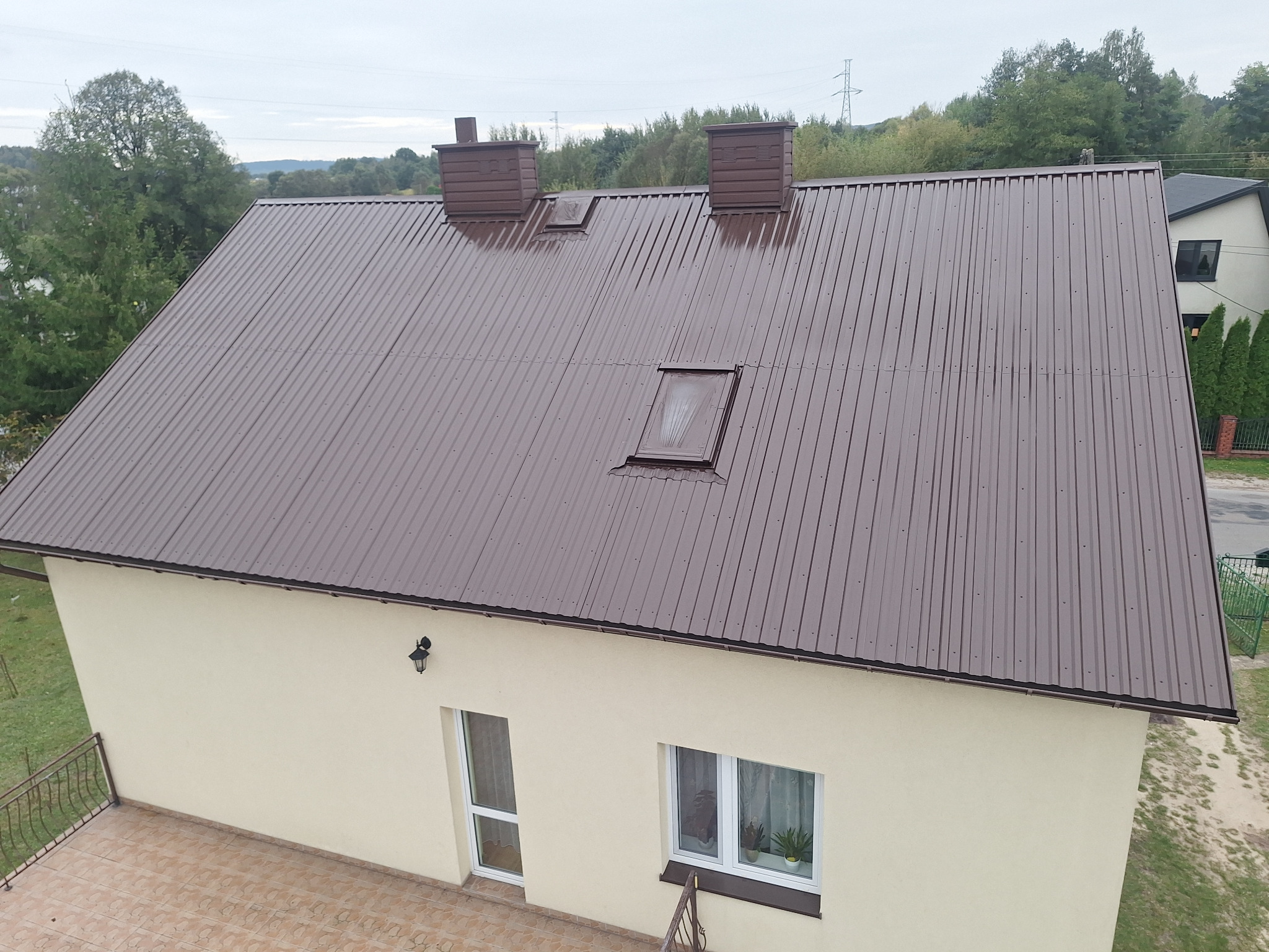 Dom jednorodzinny z brązowym dachem z blachy trapezowej, dwoma kominami i oknem dachowym, widok z góry.