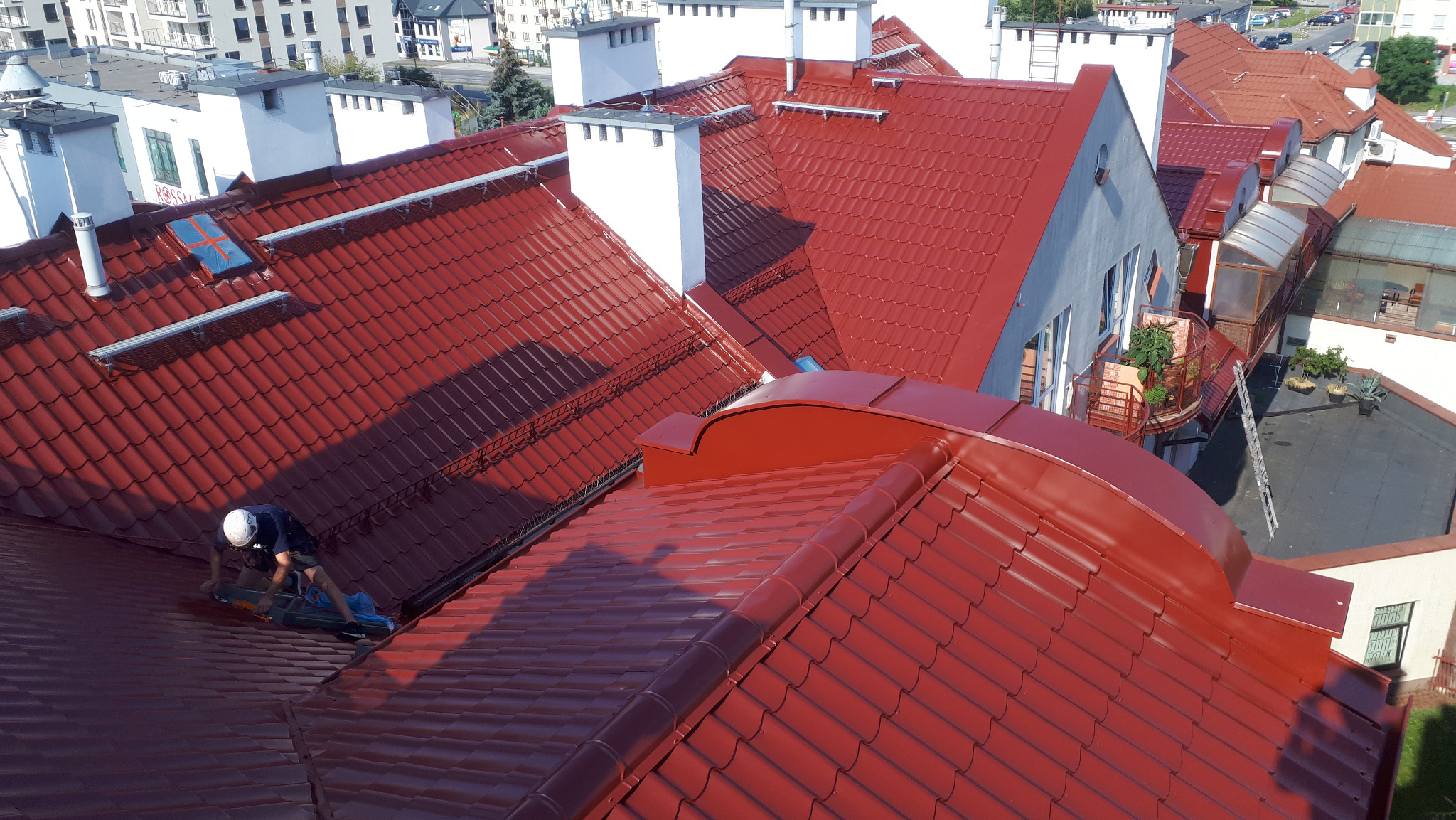 Pracownik w kasku, z narzędziami na dachu z czerwonej dachówki, widok z góry na budynki mieszkalne w tle.